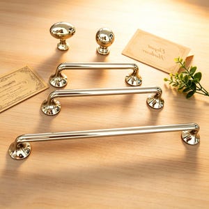 May include: A collection of gold-colored cabinet hardware, including two knobs and three different-sized bar pulls. The hardware is displayed on a light-colored wooden surface, with a small sprig of greenery and a decorative card.