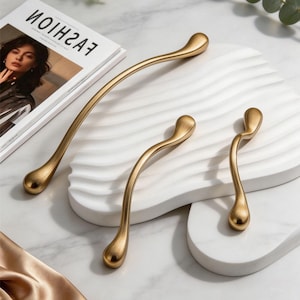May include: Four gold-toned cabinet pulls with a teardrop design. The pulls are arranged on a white, wavy surface, with a magazine and eucalyptus sprigs in the background. The pulls vary in size, with a long, curved handle and two smaller, curved handles.