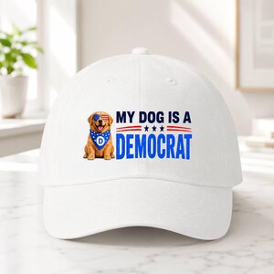 May include: A white baseball cap featuring a graphic of a golden retriever wearing American flag-patterned sunglasses and a blue bandana with a white letter D. The text reads MY DOG IS A DEMOCRAT in blue and black with red accent lines and stars.