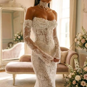 Peut inclure: Une robe longue blanche en dentelle avec des manches longues, une encolure décolletée avec un détail drapé sur le corsage et un collier ras-du-cou en dentelle assorti. Le vêtement est fait d'un tissu en dentelle florale à la coupe ajustée.
