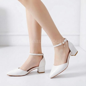 Könnte beinhalten: Weiße Pumps mit spitzer Zehenpartie und Knöchelriemen. Die Schuhe haben einen Blockabsatz und einen kleinen Anhänger am Knöchelriemen. Die Absätze sind etwa 5 cm hoch.