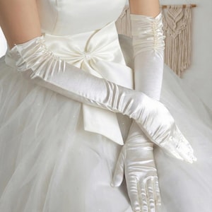 May include: Ivory satin opera gloves. These elegant, long gloves extend past the elbow and feature a ruched detail at the upper arm. The gloves are shown against a white satin gown with a large bow.