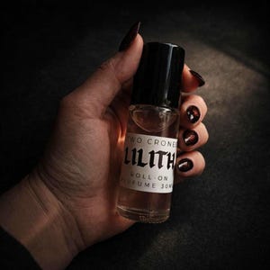 May include: A clear glass roll-on perfume bottle with a black cap. The label reads "TWO CRONES LILITH ROLL-ON PERFUME 30ml" in a gothic font. The bottle is held against a dark background.