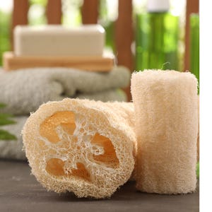 May include: Two natural loofah sponges, one cut open to show the interior, and a cylindrical one standing upright. The sponges are a light beige colour, with a textured, porous appearance. In the background, there is a bar of soap on a wooden tray and a stack of towels.
