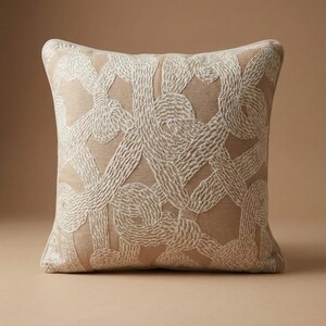 May include: Square beige throw pillow with an intricate, off-white embroidered rope design. The pillow has a neutral color palette, making it suitable for various home decor styles. The textured embroidery adds visual interest.