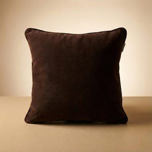 May include: A square, dark brown cushion with a soft, velvety texture and a darker brown trim. The cushion is placed on a light brown surface. The cushion is designed for decorative purposes.