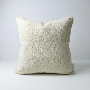 May include: A square, off-white pillow with a textured, plush surface. The pillow has a soft, inviting appearance, perfect for adding comfort and style to any living space. The neutral color allows it to complement various decor styles.