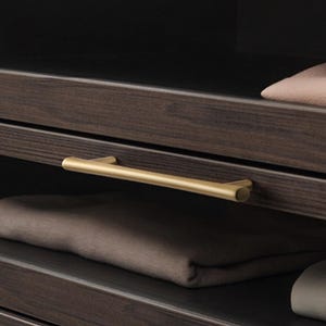 以下が含まれることがあります： ゴールドカラーの取っ手が付いた、濃い茶色の木製クローゼット。引き出しは部分的に開いており、折りたたまれた衣類が見えます。革のバッグとニット帽が上の棚に見えます。