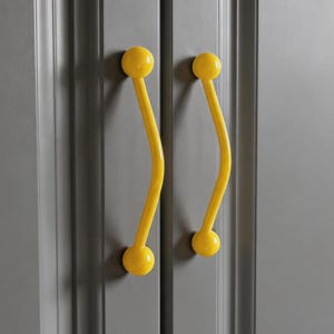 May include: Close-up of a gray cabinet with two yellow, curved handles. Each handle has a ball at the top and bottom, with a curved stem connecting them. The cabinet doors are a solid, neutral gray color.