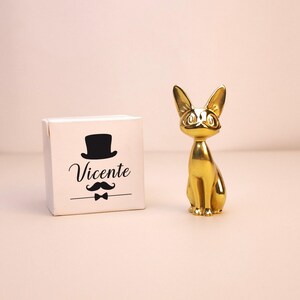 Puede incluir: Una figura de gato de color dorado brillante con orejas grandes y puntiagudas y ojos grandes, junto a una caja cuadrada blanca con un gráfico negro de un sombrero de copa, un bigote, una pajarita y el texto impreso Vicente en cursiva.