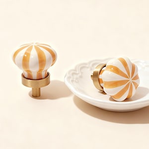 Op de afbeelding: Twee decoratieve kastknoppen met een wit en oranje gestreept ontwerp. Eén knop staat rechtop en de andere ligt op een wit, geschulpte bord. De knoppen hebben een messing voet en steel.