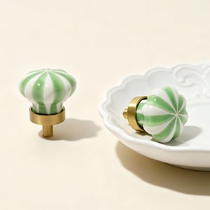 May include: Two decorative cabinet knobs. Each knob features a green and white striped design on a rounded, flower-like shape. The knobs have a brass base and stem. One knob is standing upright, and the other is resting on a white, scalloped-edge plate.