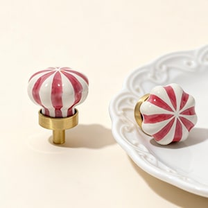 Peut inclure: Deux boutons de meuble décoratifs avec un motif à rayures rouges et blanches. Les boutons ont une forme ronde en forme de fleur et sont en céramique. La base est en métal doré. Un bouton est droit et l'autre est posé sur une assiette blanche.