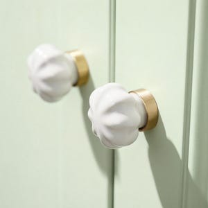 May include: Close-up of two white ceramic cabinet knobs with a fluted design, mounted on a light green cabinet door. The knobs have a gold-colored metal base. The cabinet door is vertically paneled.