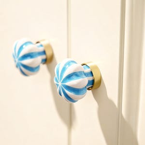 May include: Two decorative cabinet knobs. Each knob features a white and blue striped design with a gold-colored base. The knobs are mounted on a white surface, offering a pop of color and style.