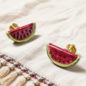 May include: Two watermelon slice cabinet knobs with brass hardware. The knobs are red with black seeds and green rind. They are set against a white fabric background with a decorative trim.