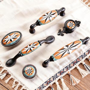 May include: A selection of decorative cabinet hardware with a starburst design in orange, white, and black. The hardware includes handles, knobs, and pulls with a turquoise accent and dark bronze finish.