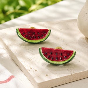 May include: Two drawer knobs shaped like watermelon slices. Each knob features a bright red fruit interior with black seeds, a white rind, and a dark green outer edge. The hardware is finished with a metallic gold-tone circular base.