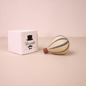 Puede incluir: Una caja cuadrada blanca con un gráfico negro de un sombrero de copa, un bigote, una pajarita y el nombre Vicente, junto a un adorno de globo aerostático color crema con rayas verticales gris oscuro y una base marrón.