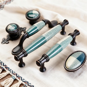 Puede incluir: Un conjunto de herrajes para armarios con tiradores y pomos. Los tiradores tienen un patrón de cuadros verdes y blancos con bases de bronce oscuro. Los pomos y tiradores tienen un diseño a juego. Los herrajes están dispuestos sobre un paño blanco.