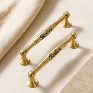 May include: Two gold-toned metal cabinet pulls with a smooth, cylindrical design and rounded ends. Each handle features the word Pandora engraved in a black, cursive script along the center of the bar, supported by two circular bases.