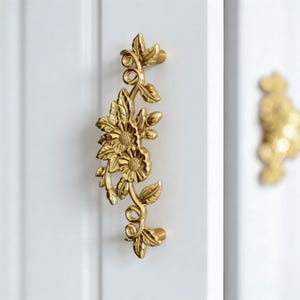 May include: A gold-coloured floral door handle featuring a detailed design of flowers and leaves. The handle is mounted on a white door, highlighting its intricate craftsmanship. The handle's design includes two flower heads and several leaves.