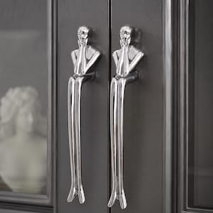 May include: Two polished chrome cabinet handles. Each handle features a stylized figure with hands near the face, atop a long, slender pull. The handles are mounted on a dark gray cabinet door. A bust is visible through the glass.