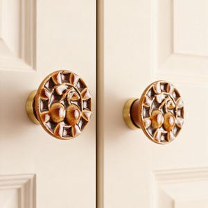 Puede incluir: Dos tiradores decorativos para armarios con diseño de cerezas. Los tiradores son redondos con una cara de cerámica marrón y crema y una base de latón. El diseño de cerezas es en relieve y detallado, añadiendo un toque de elegancia.