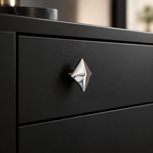 May include: A diamond-shaped, polished silver metal cabinet knob with a faceted surface and a small engraved logo, mounted on a matte black drawer front. The knob features sharp, geometric edges and a reflective finish that contrasts with the dark surface.