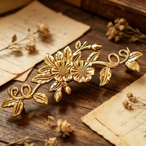 Puede incluir: Un tirador de armario floral dorado con dos flores grandes y hojas. El herraje decorativo es de metal y está diseñado para montarse en un armario o cajón. El tirador se muestra sobre una superficie de madera con papel vintage.