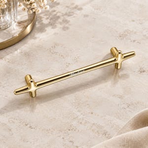 Peut inclure: Une poignée de meuble en métal doré poli dotée d'une fine barre horizontale aux extrémités en forme de croix. Le centre de la barre présente le texte gravé "Pandora" dans une police cursive délicate, reflétant la lumière sur sa surface lisse.