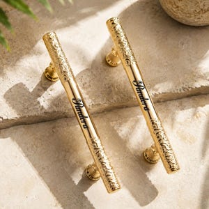 May include: Two gold-toned metal cabinet handles featuring a cylindrical shape with ornate, textured ends and a smooth center section. Each handle is engraved with the word Pandora in a black script font and includes two circular mounting bases.