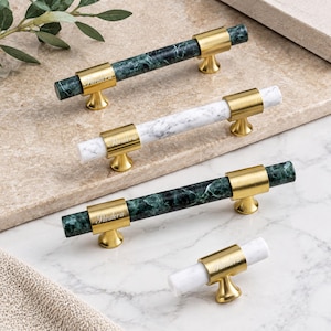 May include: Four cabinet pulls featuring cylindrical stone bars in dark green or white marble with gold-toned metal mounts. Each mount is engraved with the word Pandora. The set includes three long horizontal handles and one smaller knob.