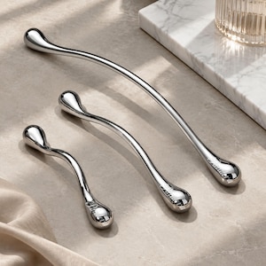 May include: Three curved, silver-toned metal handles of varying lengths, each featuring a smooth, polished finish and a teardrop-shaped end. The handles are arranged diagonally, with the longest at the top and the shortest at the bottom.