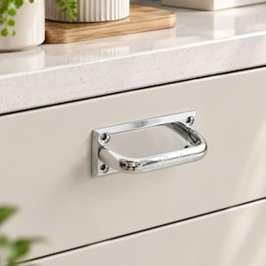 May include: A polished chrome drawer pull with a rectangular mounting plate featuring four screw holes. The handle has a curved, U-shaped design and is mounted horizontally on a smooth, light beige cabinet drawer front with a reflective metallic finish.