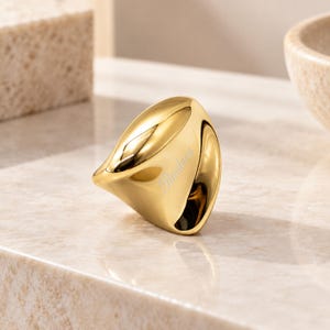 May include: A polished gold-tone ring with a smooth, organic, and asymmetrical shape. The surface of the ring features the engraved word Pandora in a cursive script, reflecting light across its curved, reflective, and metallic exterior finish.