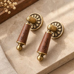 May include: Two drop-style cabinet pulls featuring a dark wood handle with the word Pandora engraved in script, accented by antique brass metal hardware with a circular base plate displaying a detailed floral petal pattern design.