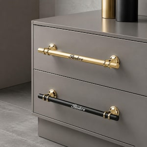 May include: Two horizontal drawer pulls mounted on a grey cabinet. The top pull is polished gold with the word Pandera engraved in script. The bottom pull is matte black with gold accents and the word Pandera engraved in script on the centre.