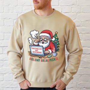 May include: Beige sweatshirt with a graphic of Santa Claus wearing glasses, a red hat, and suit, looking at a laptop. The laptop screen reads "CHRISTMAS.EXE NOT RESPONDING". Below the laptop is the text "HOLIDAY BREAK PLEASE".
