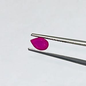 May include: A vibrant, pear-shaped, fuchsia gemstone is held by a pair of silver tweezers against a white background. The gemstone's polished surface reflects light, showcasing its smooth, rounded form. This image highlights the gemstone's color and shape.