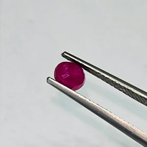 May include: A faceted, oval-shaped ruby gemstone held by a pair of silver tweezers. The ruby is a deep, saturated red color, with visible facets that catch the light. The background is a plain, light gray.