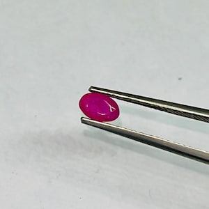 Può includere: Una vibrante gemma di rubino di forma ovale, tenuta da pinzette d'argento. Il rubino mostra un colore rosa intenso e saturo, con una superficie liscia e lucida. Le pinzette sono posizionate su uno sfondo chiaro e uniforme.