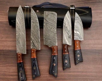 May include: A set of five Damascus steel knives with wooden and black handles, displayed with a black leather knife roll. The blades have a unique patterned design. The handles are a combination of wood and black material.