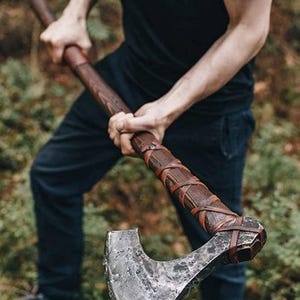 May include: Axe with a wooden handle and a metal blade. The handle is wrapped with brown leather straps. The axe is held by a person wearing a black shirt and dark blue pants. The background is a forest.