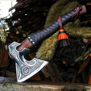 May include: A hand axe with a polished silver-tone metal head featuring intricate engraved patterns and a central triangular knot symbol. The wooden handle is wrapped in dark brown leather with a braided texture and a decorative orange and brown tassel.