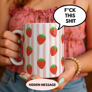 May include: White ceramic mug with a repeating pattern of red strawberries and green leaves against pink and white stripes. A speech bubble with the text "F*CK THIS SHIT" is on the right. The bottom of the mug has the text "HIDDEN MESSAGE".