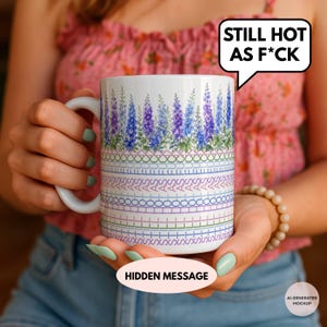 May include: A white ceramic mug featuring a top band of blue and purple lupine flowers. Below the flowers, the mug displays a repeating pattern of the text STILL HOT AS F*CK in a cross-stitch style, followed by rows of geometric cross-stitch symbols.