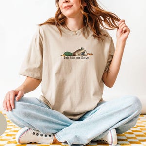 Pode incluir: Uma t-shirt de manga curta na cor areia com um gr&aacute;fico de um pato-real deitado de barriga para baixo. Abaixo do pato, o texto Ich bin am Ente aparece numa fonte preta simples. A t-shirt tem gola redonda e um corte descontra&iacute;do.