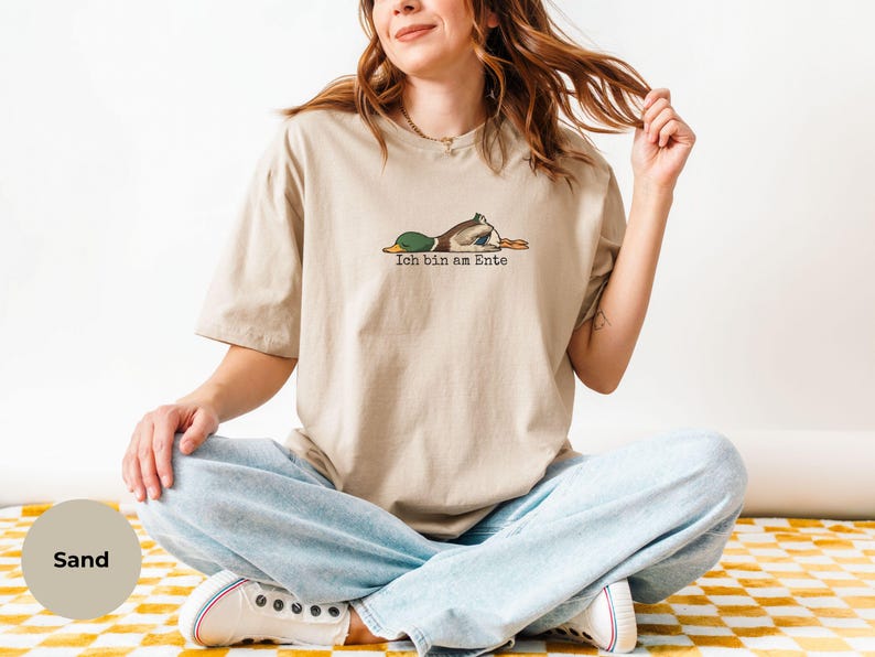 Pode incluir: Uma t-shirt de manga curta na cor areia com um gr&aacute;fico de um pato-real deitado de barriga para baixo. Abaixo do pato, o texto Ich bin am Ente aparece numa fonte preta simples. A t-shirt tem gola redonda e um corte descontra&iacute;do.