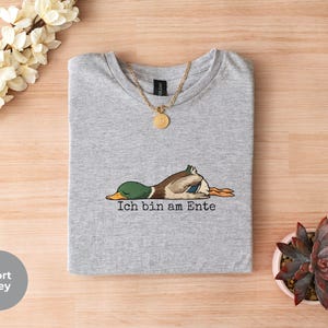 Pode incluir: Uma t-shirt cinzento desportivo com um gr&aacute;fico de um pato-real deitado de barriga para baixo e o texto Ich bin am Ente impresso a preto por baixo do pato. Um colar de corrente dourada repousa sobre o decote da t-shirt.